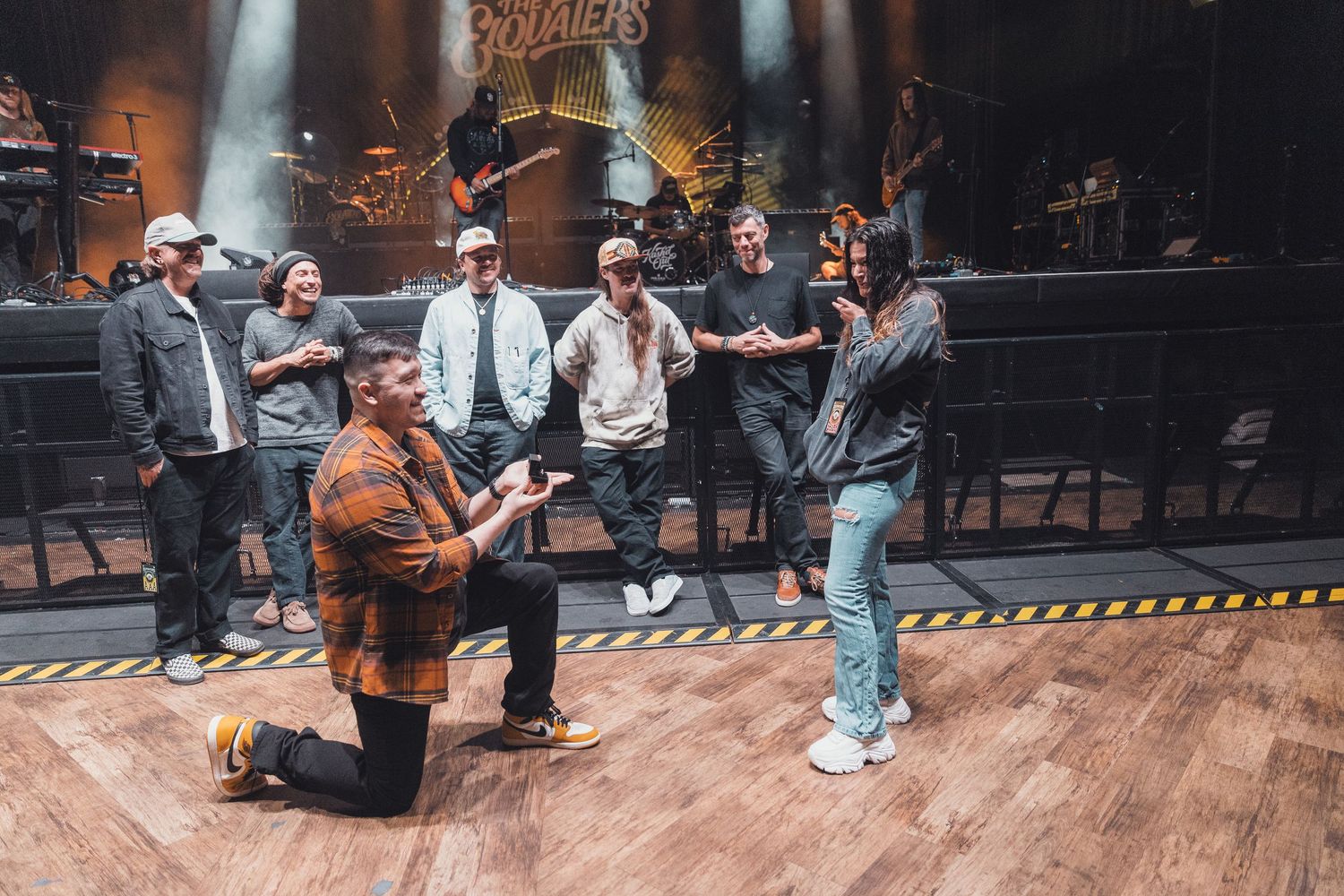 Man proposes on one knee to woman on stage at a concert venue with band members watching.