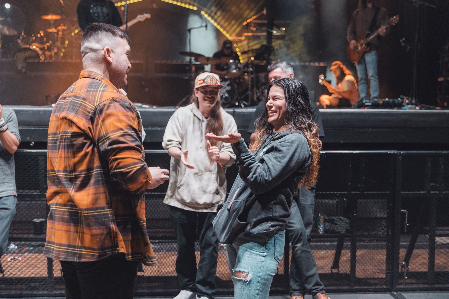 People socializing at an outdoor concert venue with a band performing on stage in the background.