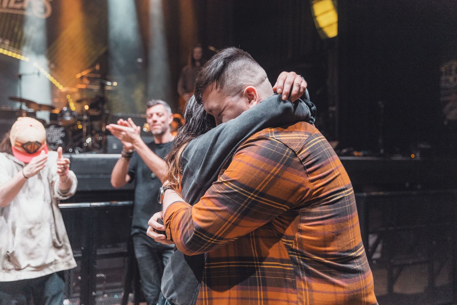 Two people share an emotional hug on a concert stage with band members and bright lights in the background.