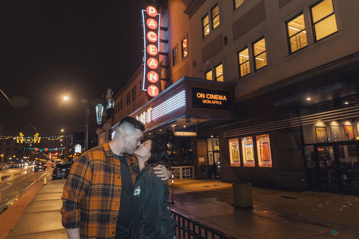 Couple sharing a romantic kiss outside a lit cinema marquee at night on a city street.