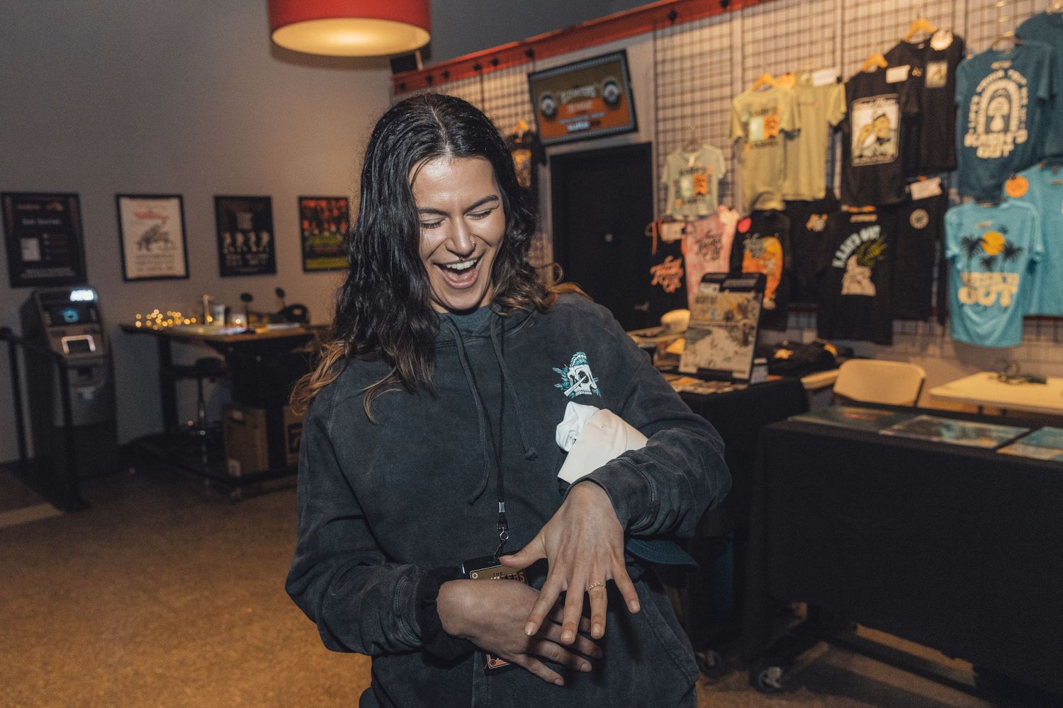 Woman laughing in a skateboard shop, wearing a dark jacket and holding a white cloth indoors.
