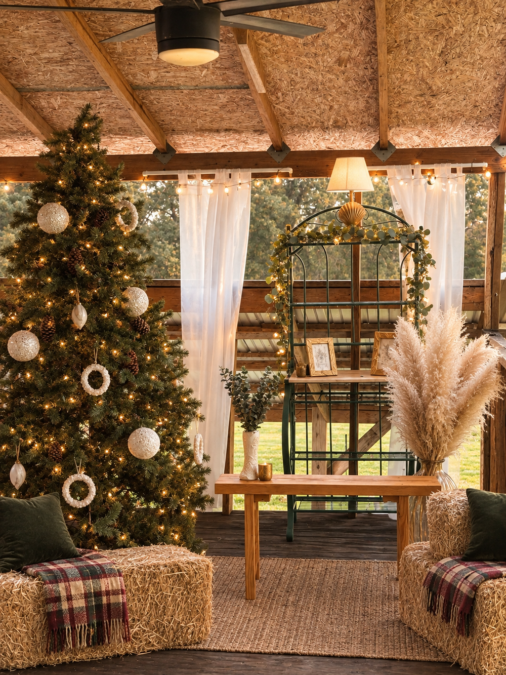Rustic Christmas barn decorated with lit tree, hay bale seating, plaid blankets, pampas grass, and sheer curtains.