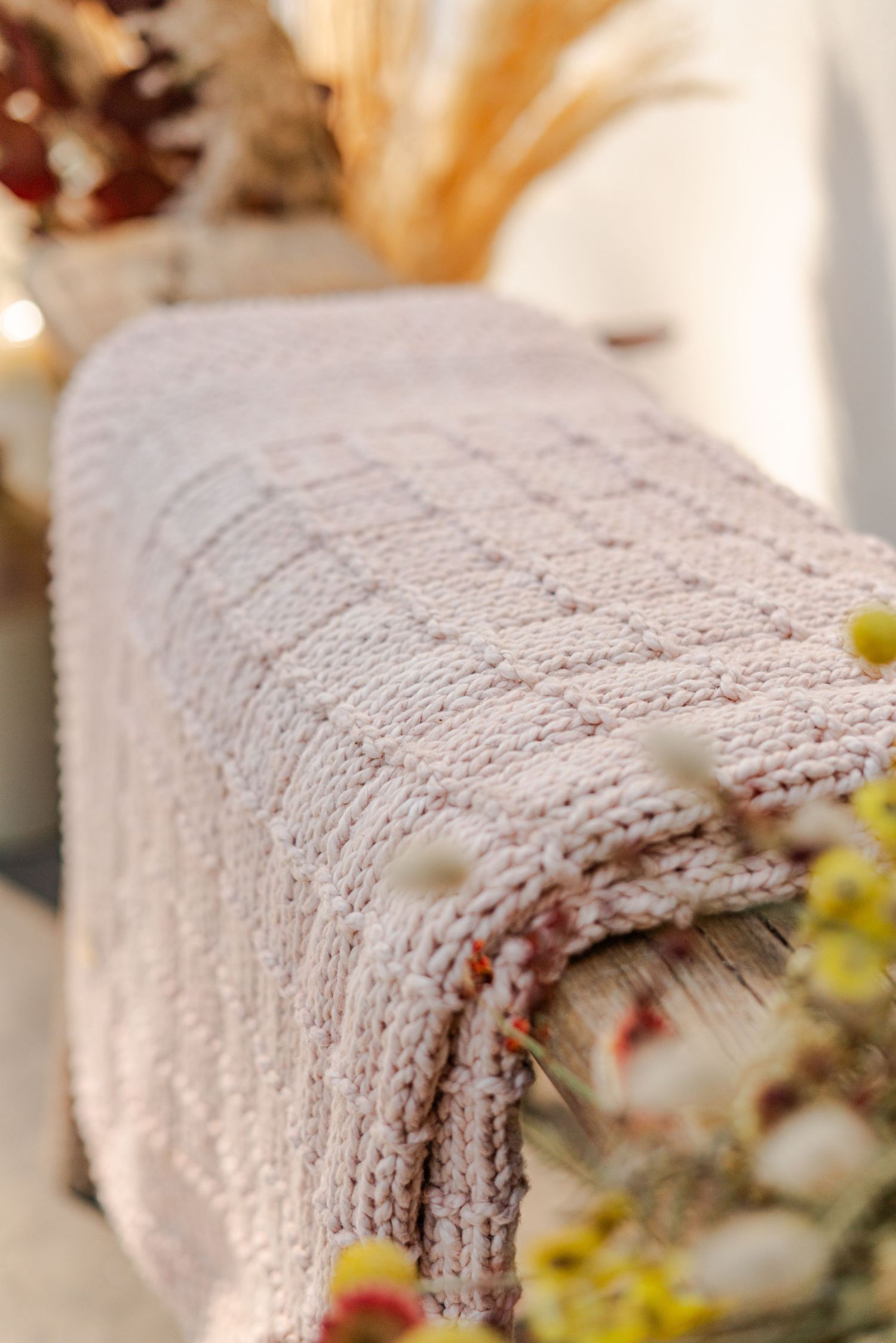 Close up detailed photography of a soft pink knitted blanket draped over a surface, surrounded by dried flowers and warm autumn decor.