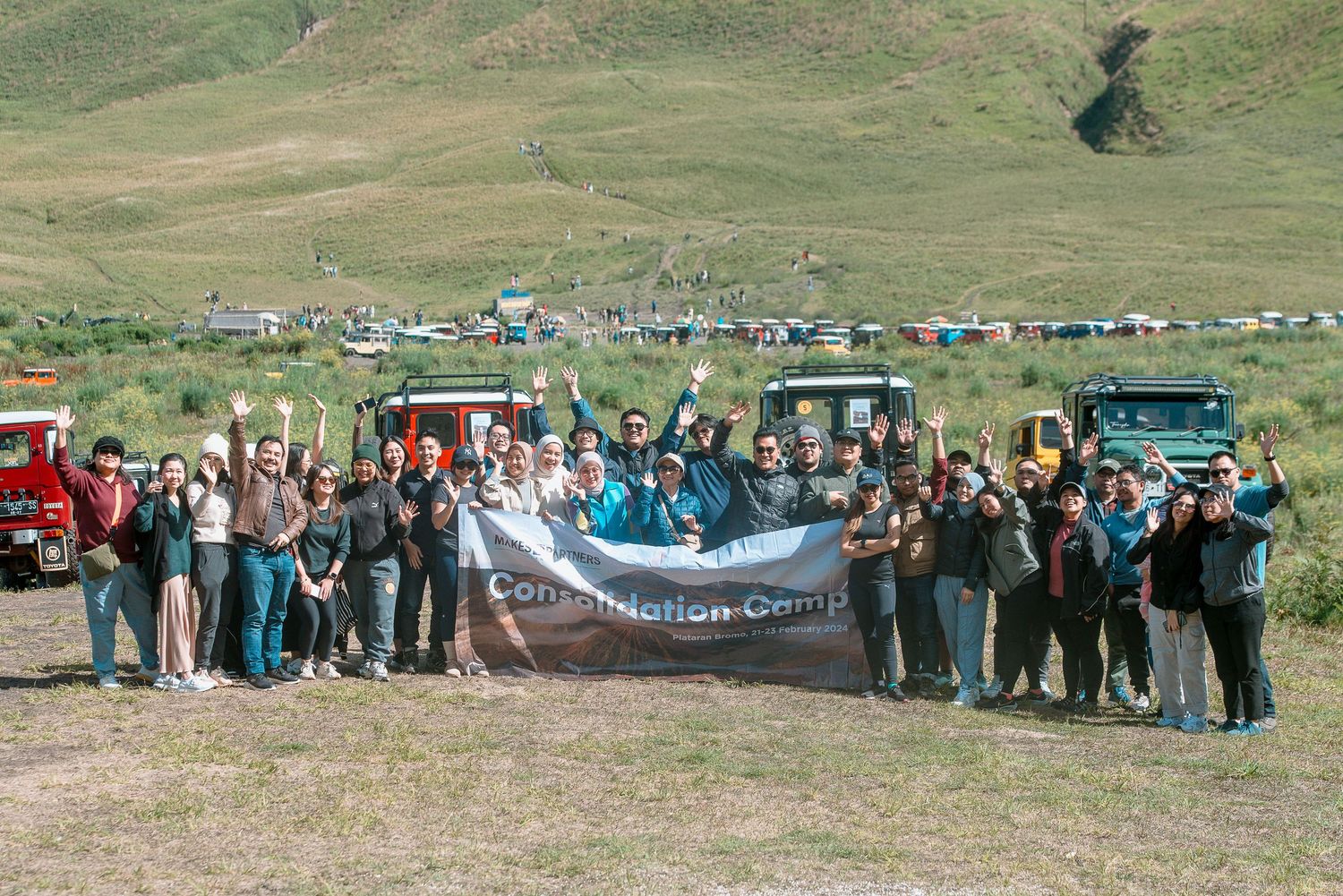 Large group of people posing outdoors holding a Compilation Camp banner in a scenic green mountain valley.