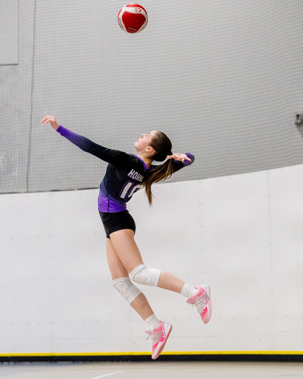 Female volleyball player jumping to serve a red and white Mikasa ball indoors.