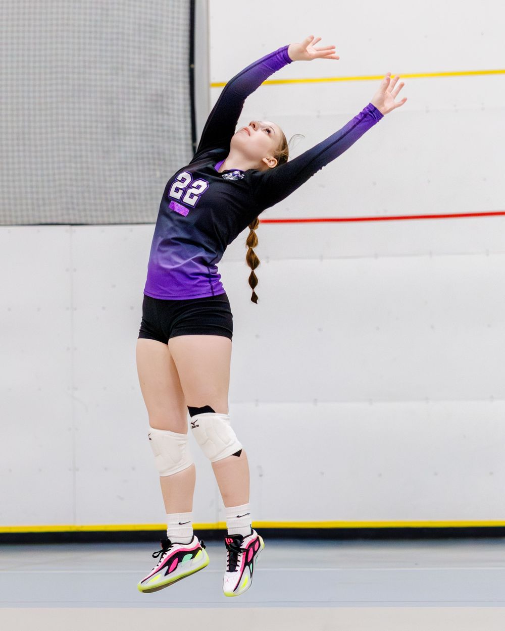 Female volleyball player number 22 in black and purple uniform jumping to serve indoors.