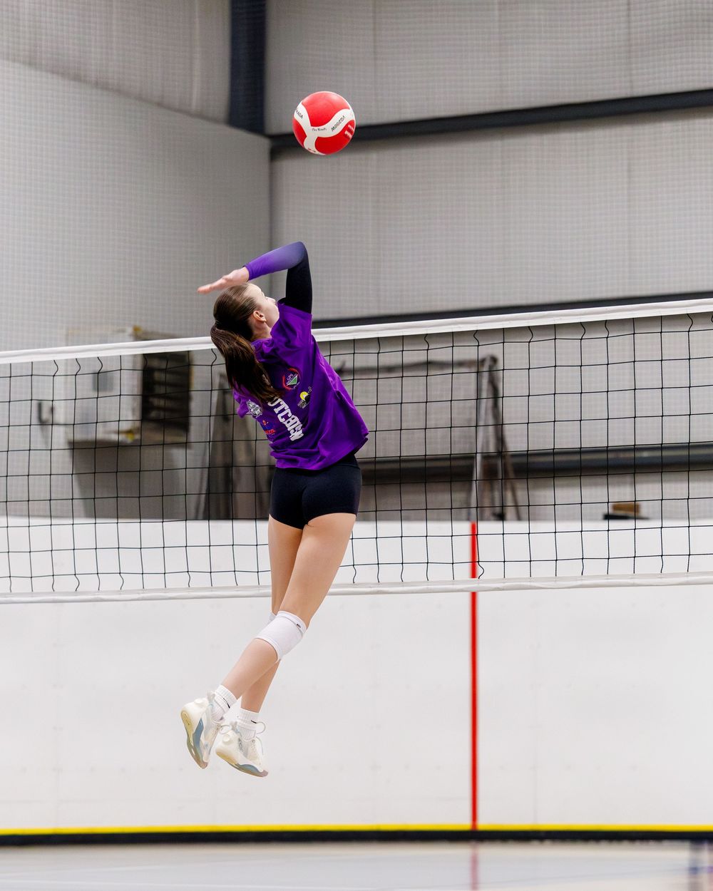 Female volleyball player in purple jersey jumping to spike the ball over the net indoors.