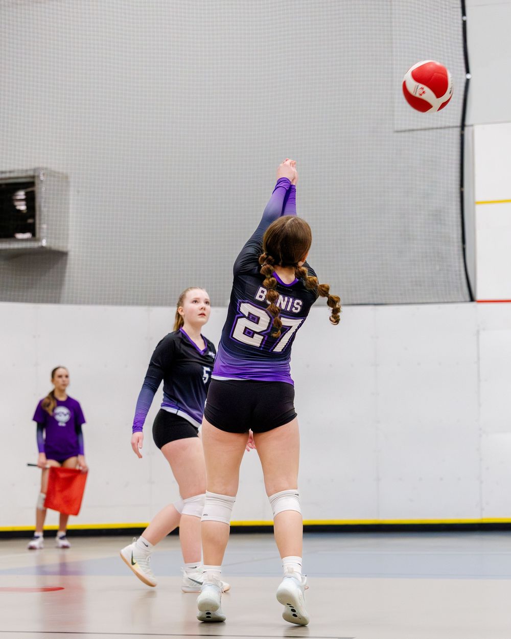 Female volleyball player number 27 serving the ball during an indoor volleyball match.