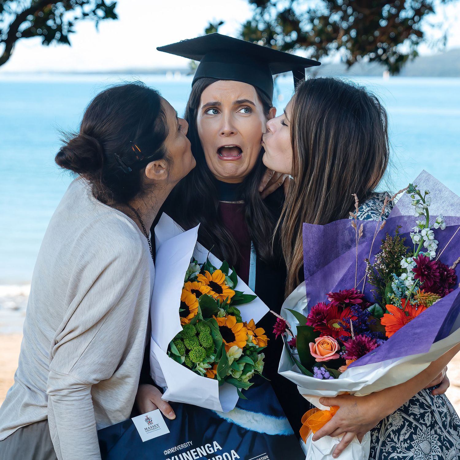 Female graduate pulling a silly face while being kissed on both cheeks and holding graduation flowers