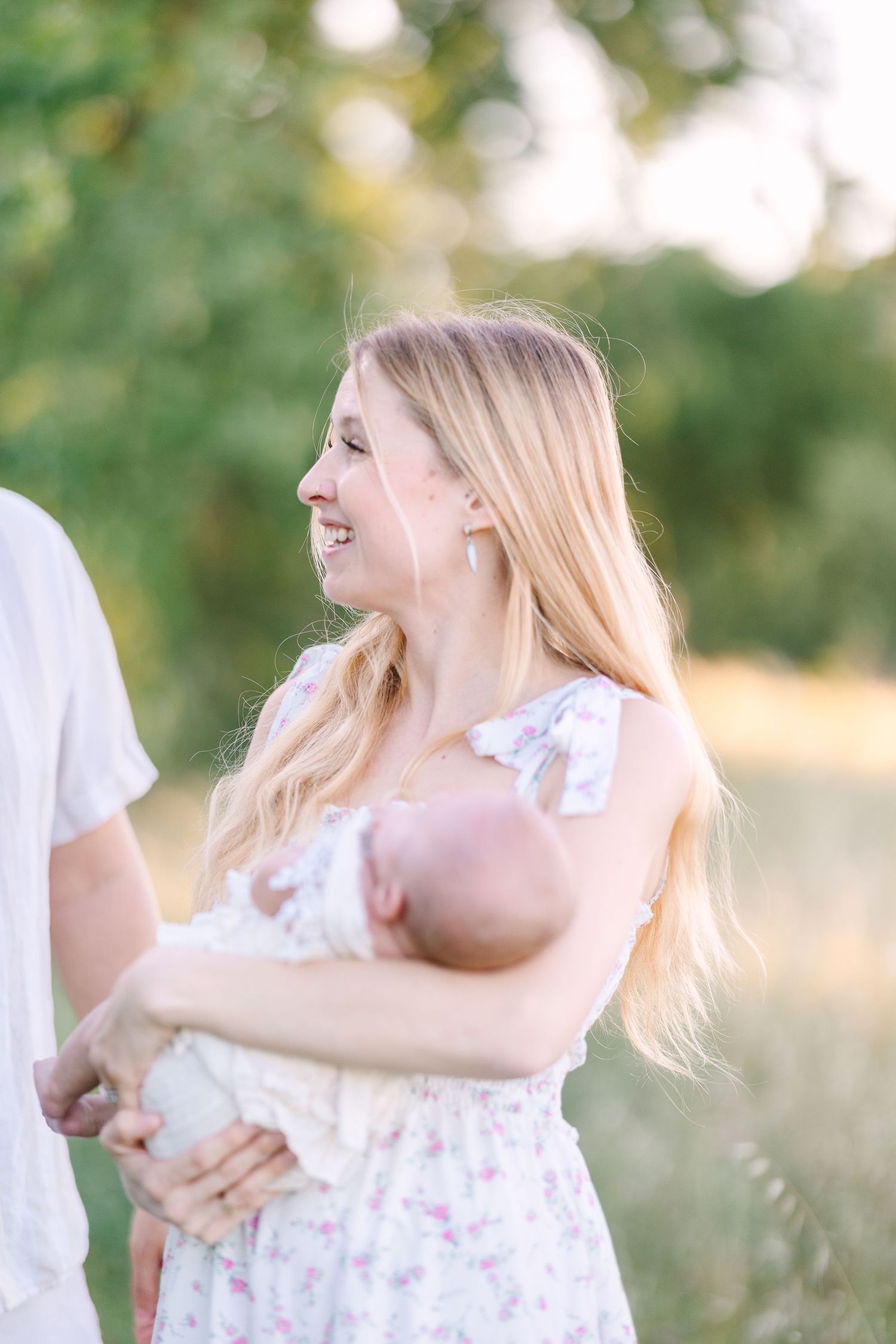 Smiling blonde mother in floral dress holds newborn baby outdoors in a sunlit green field.