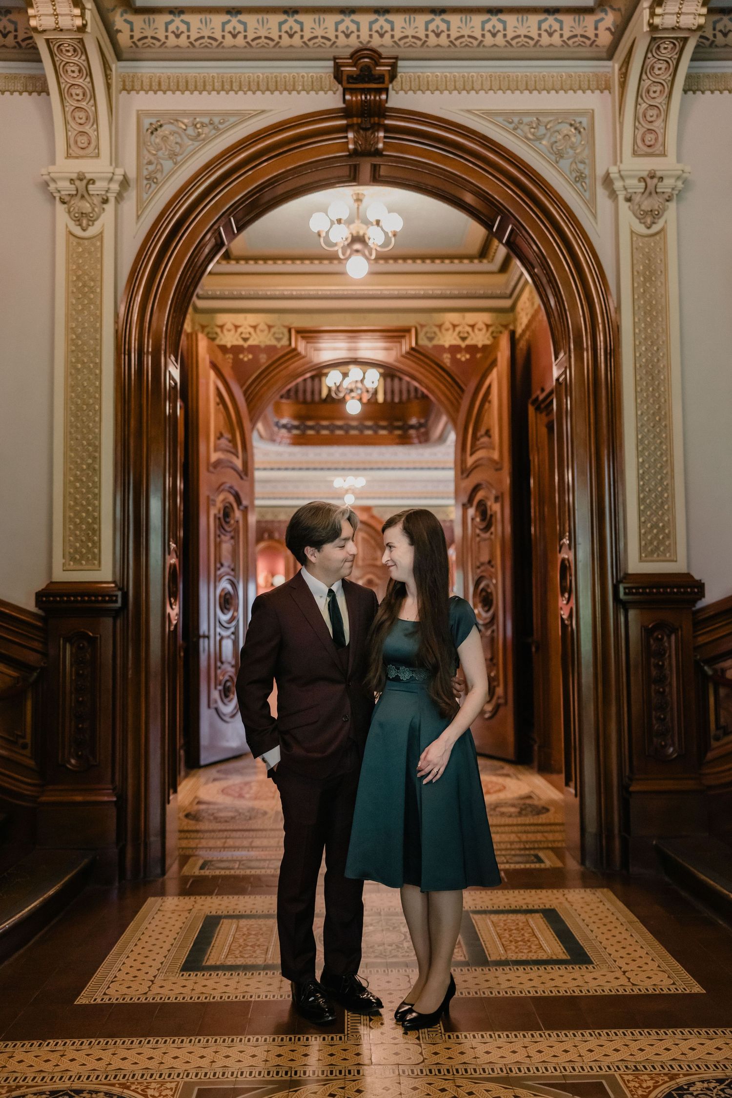 Couple in formal attire standing under ornate wooden arch in elegant historic building with chandelier.