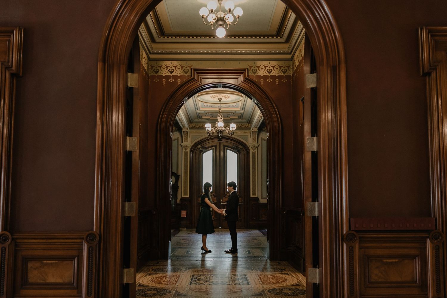 Couple holding hands in an elegant wood-paneled hallway with arched doorways and chandelier lighting.