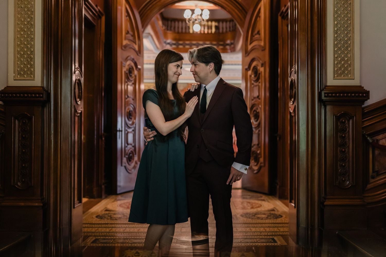 Couple in elegant attire sharing a romantic moment in an ornate hallway with chandeliers.