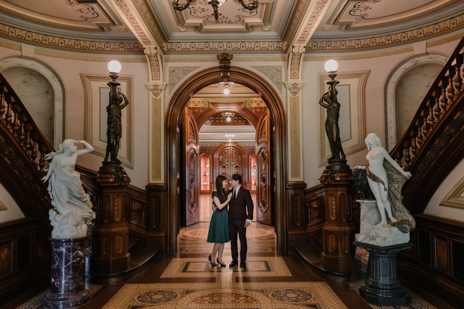Couple embracing in ornate grand hall with marble statues, gilded ceilings, and elegant staircase in historic building.