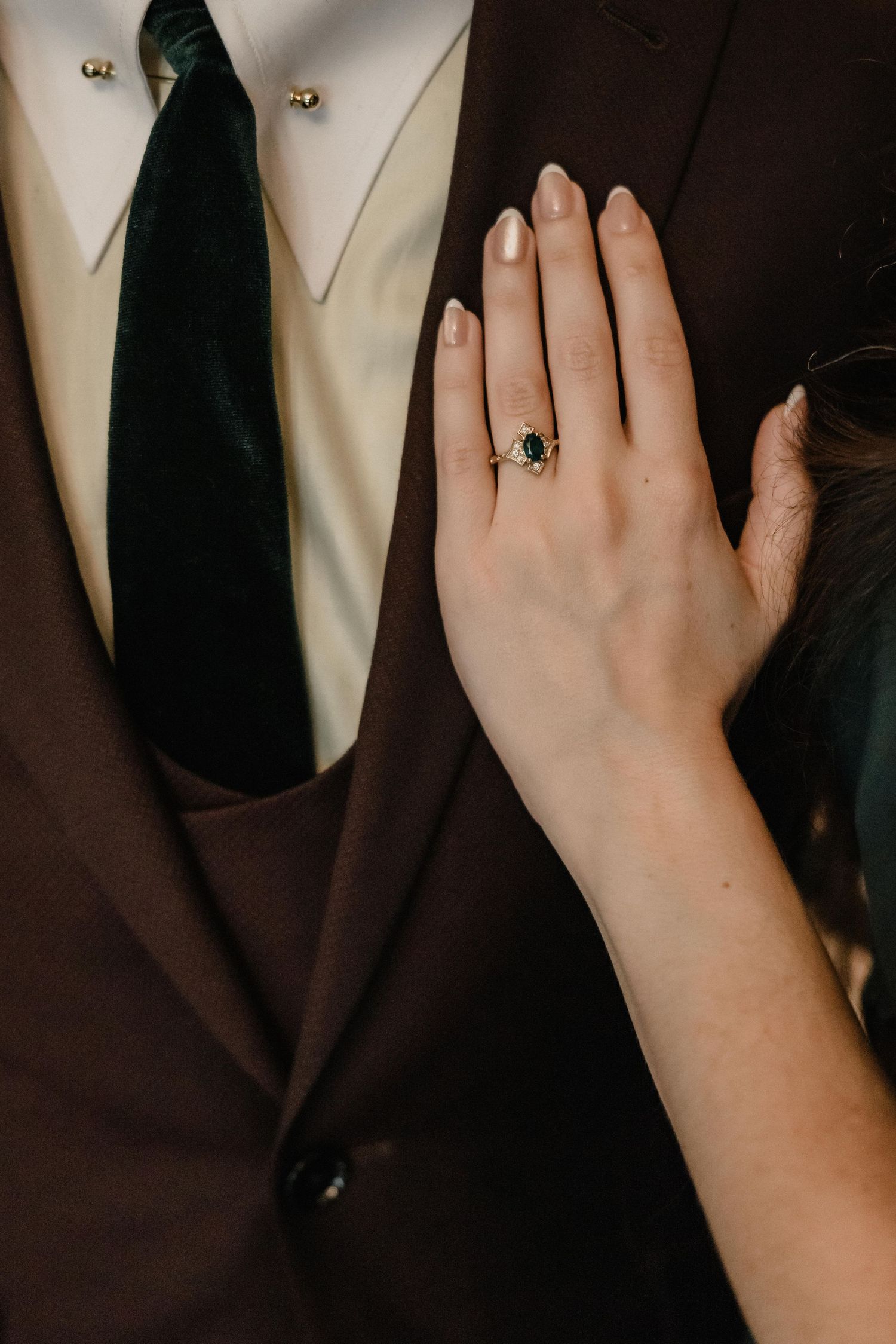 Close-up of a hand with a dark sapphire engagement ring resting on a brown suit jacket with green velvet tie.