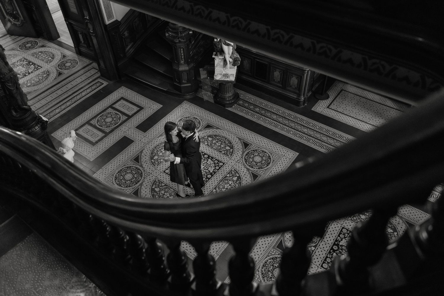 Black and white aerial view of couple embracing on ornate tiled floor beneath grand staircase.