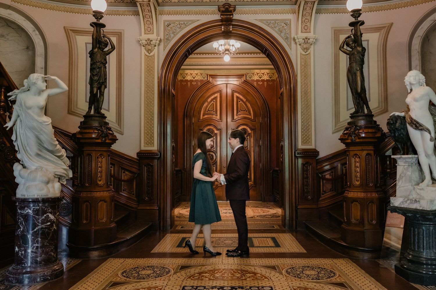 Couple holding hands in an ornate historic building hallway with marble statues and golden archways.