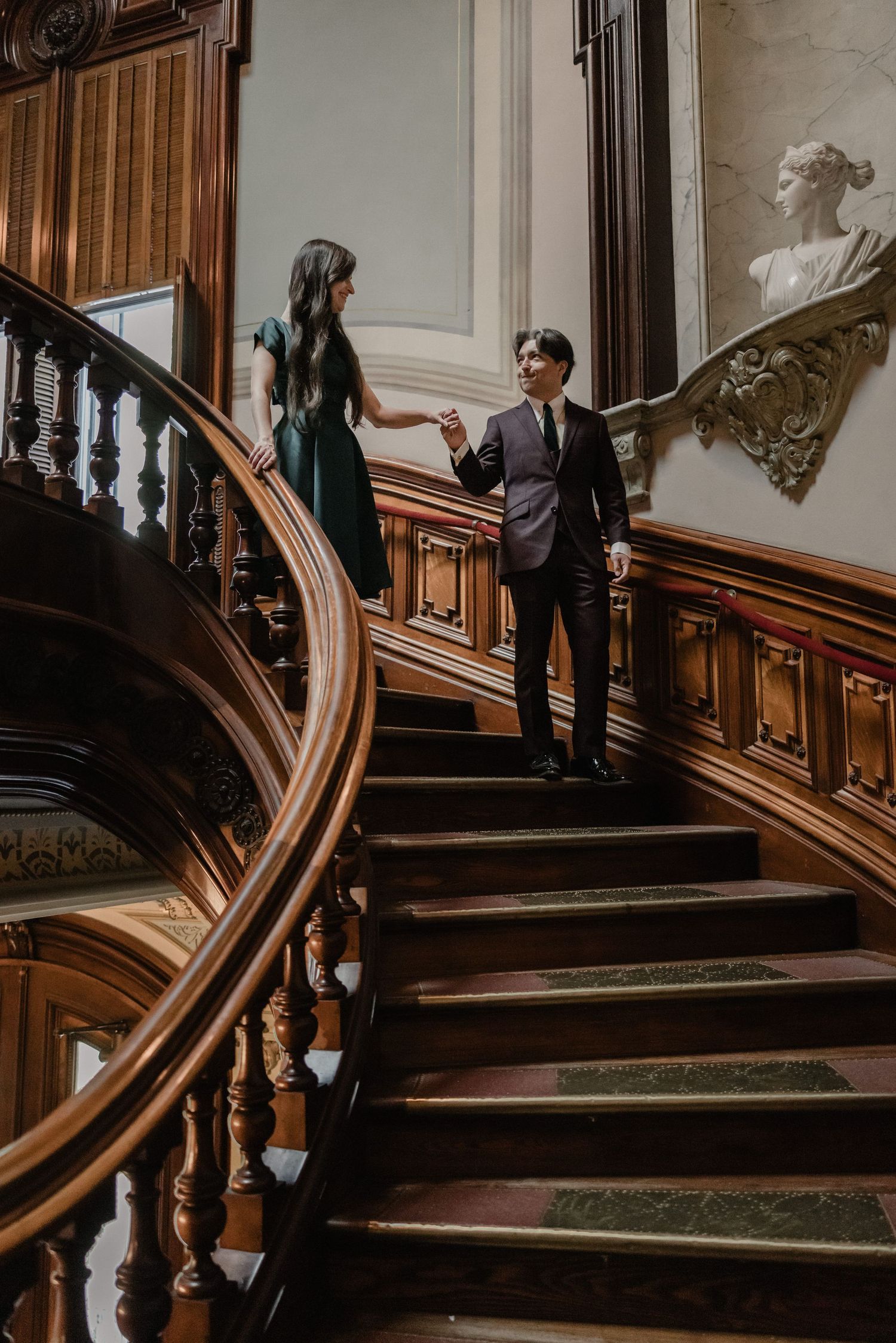 Elegant couple holding hands on ornate wooden staircase in a grand historic building with classical decor.