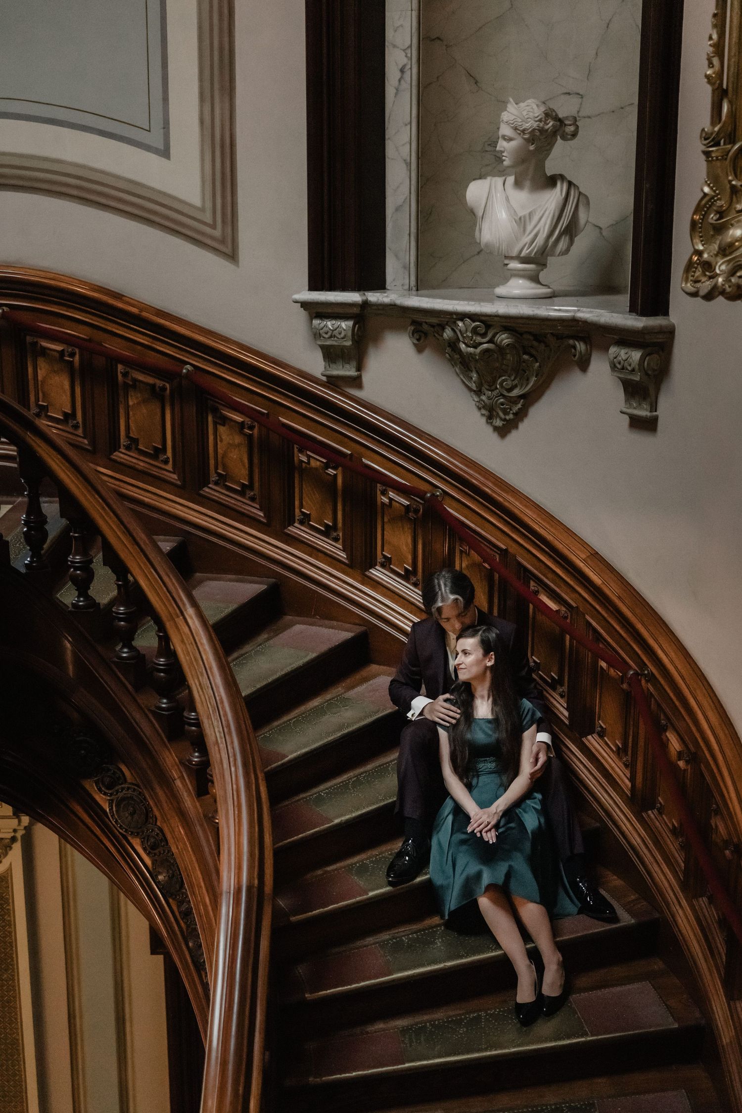 A woman in a teal dress sits on ornate wooden stairs beside a man in a historic mansion with a marble bust above.