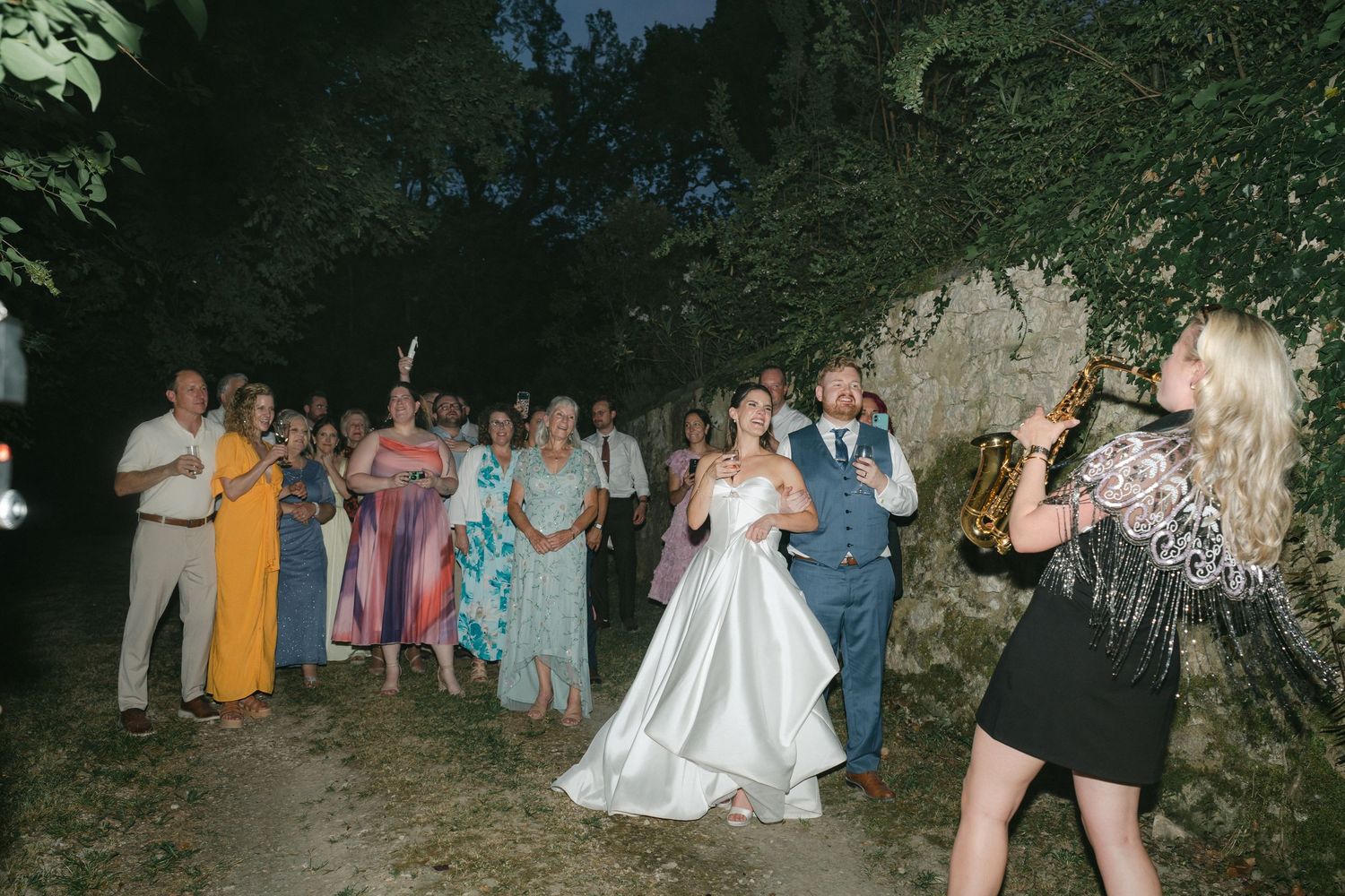 Bride and groom dancing outdoors at night while a saxophonist performs for wedding guests gathered around them.