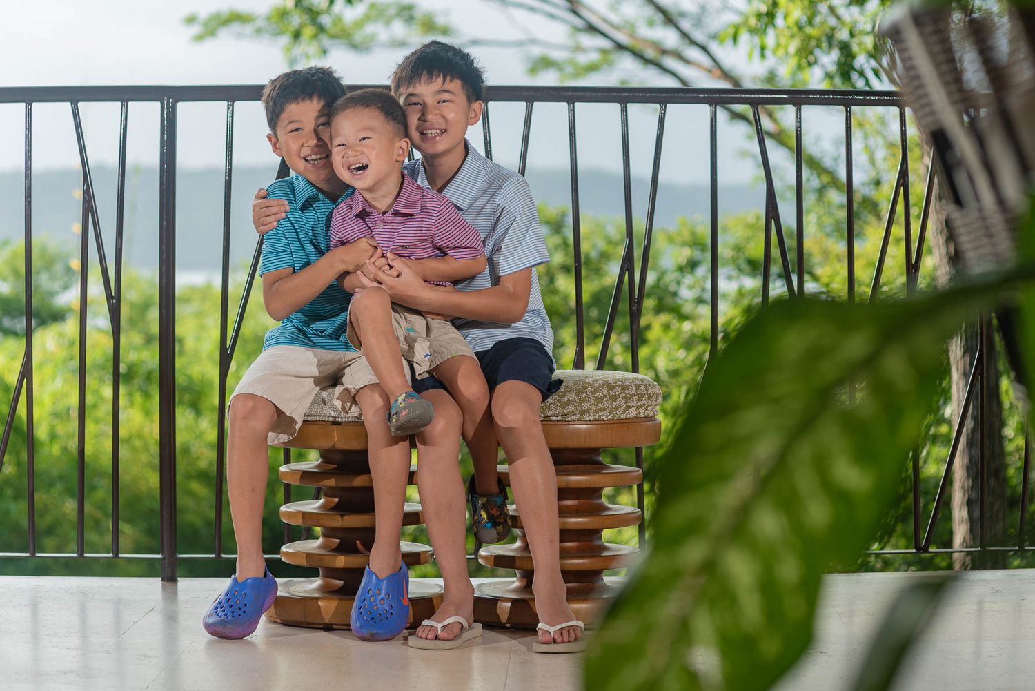 Three young boys laughing and hugging on a balcony bench with lush green trees in the background.