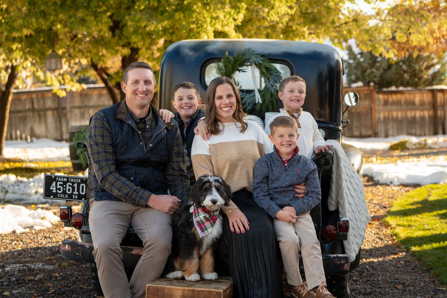 A family of six poses together leaning against a vintage black truck in front of a brick home outdoors.