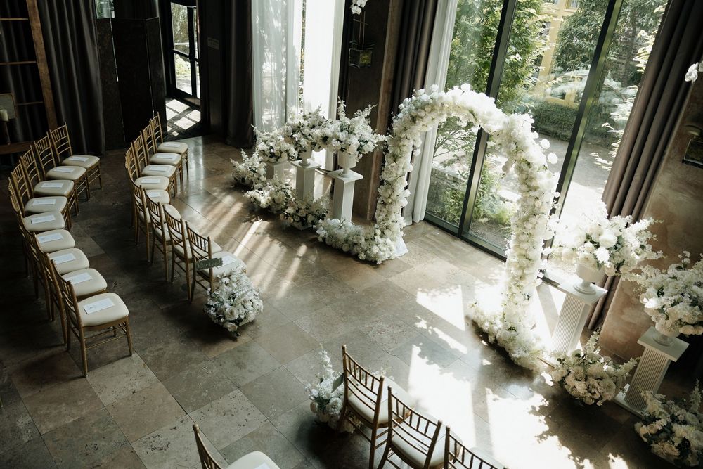 Elegant indoor wedding ceremony setup with white floral arch, chiavari chairs, and floor-to-ceiling windows.
