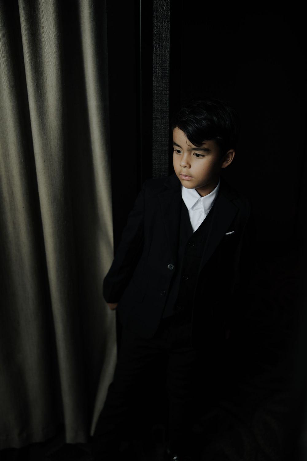 Young boy in black suit stands beside a curtain in dramatic dark studio lighting.