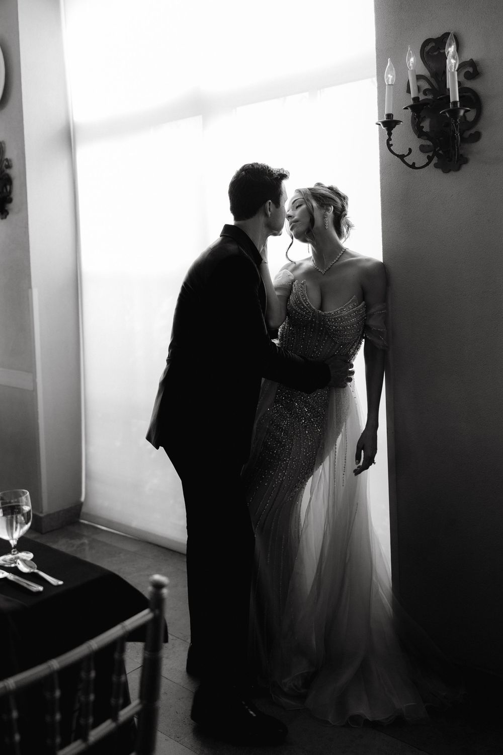 Black and white photo of a couple sharing a romantic moment, bride in beaded gown and groom in suit near a window.