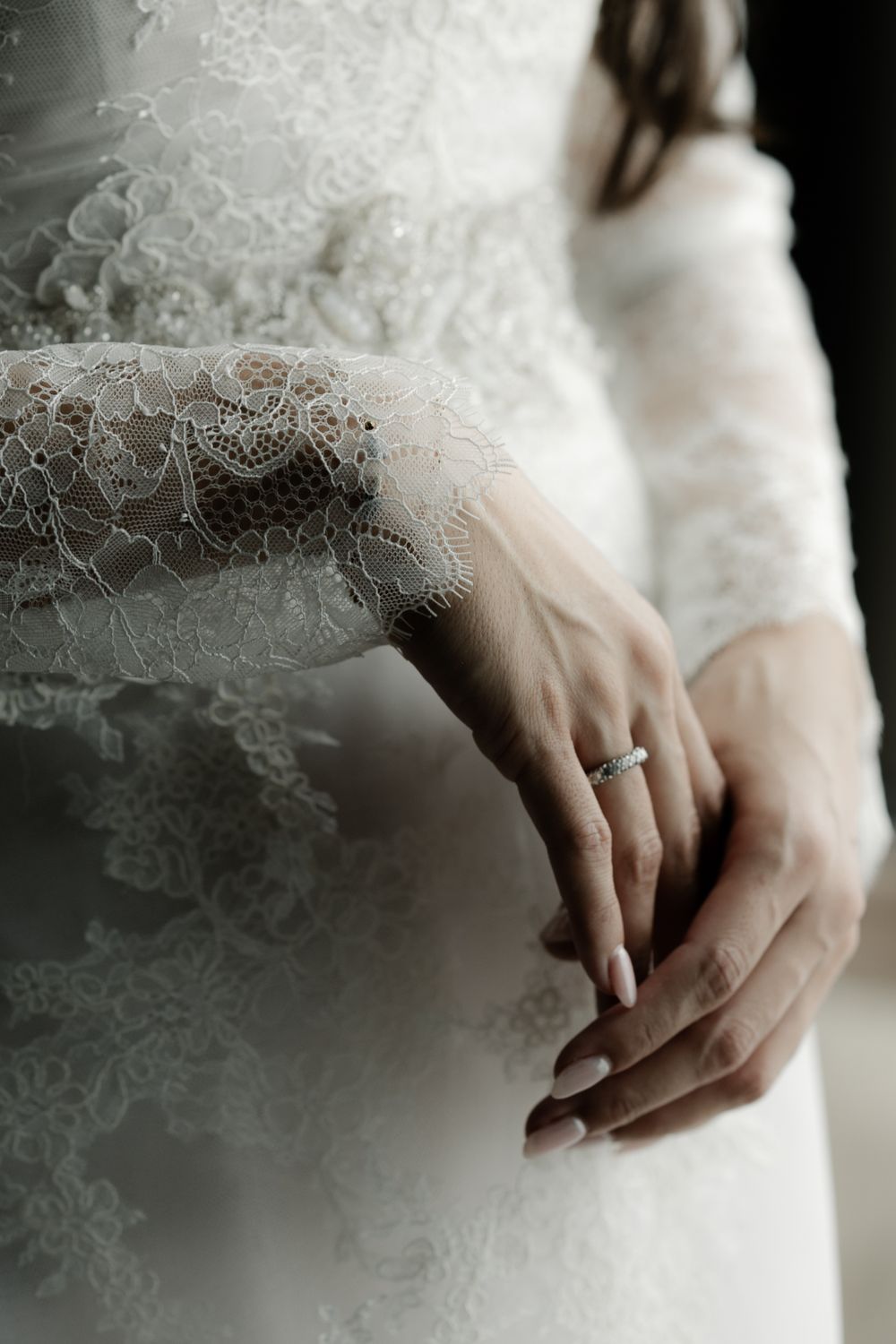 Bride in lace wedding gown with long sleeves, hands clasped showing diamond engagement ring.