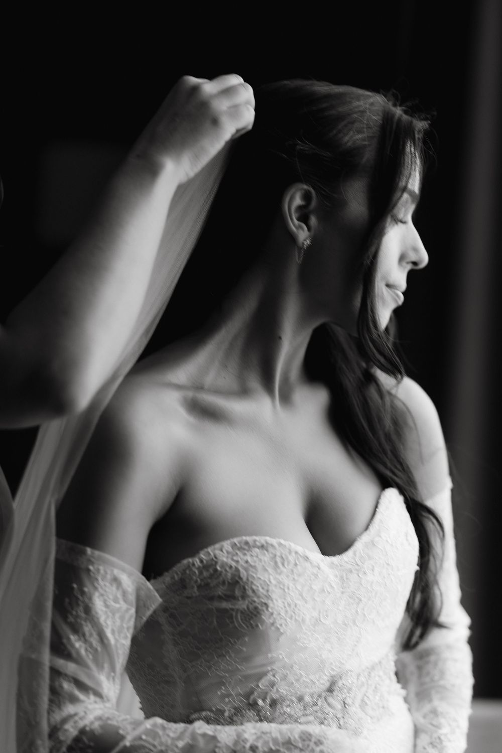 Black and white photo of a bride in a strapless lace wedding dress adjusting her veil, smiling softly.