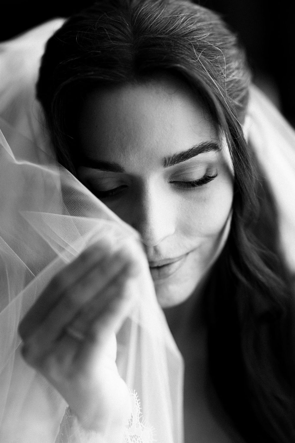 Black and white portrait of a bride holding her veil, eyes closed, with elegant makeup and flowing dark hair.