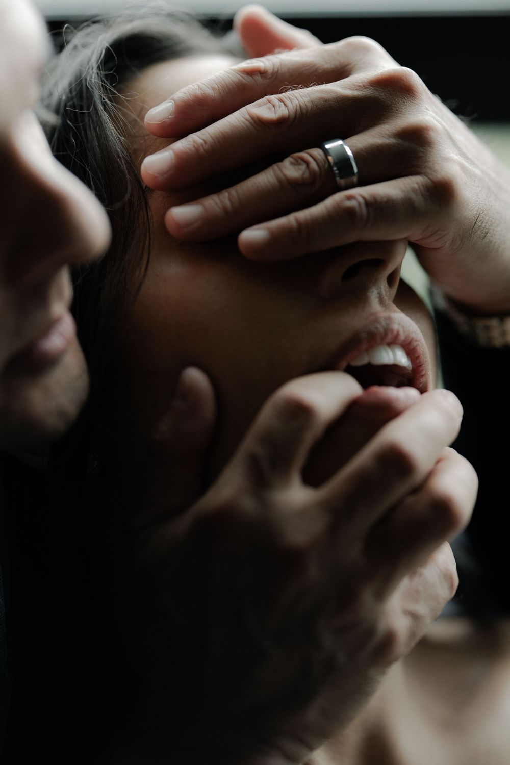 A person covering another's eyes with one hand while touching their chin, wearing a silver ring, in dramatic lighting.