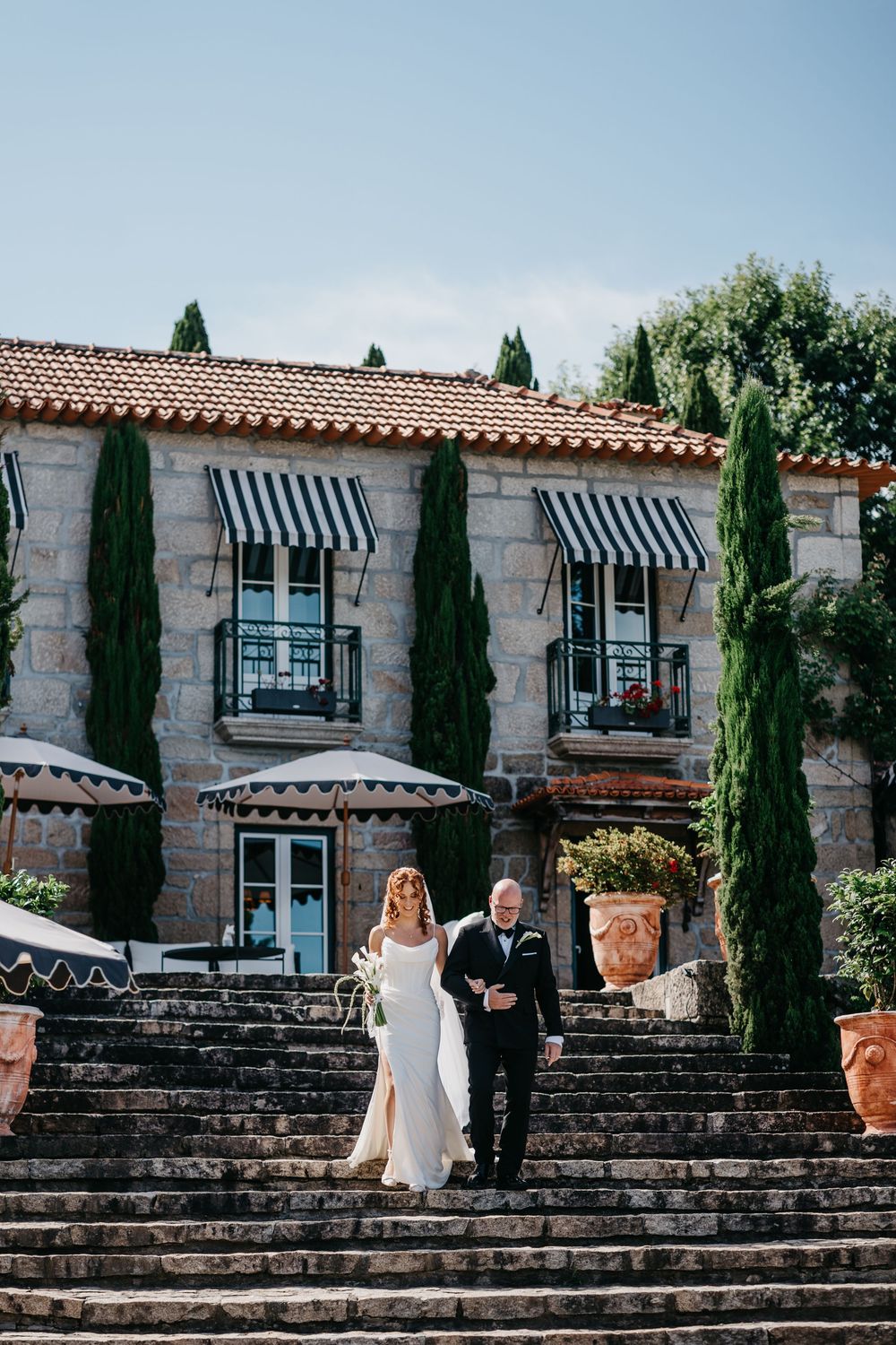 Bride and groom walk down stone steps at elegant Mediterranean villa wedding venue with cypress trees.