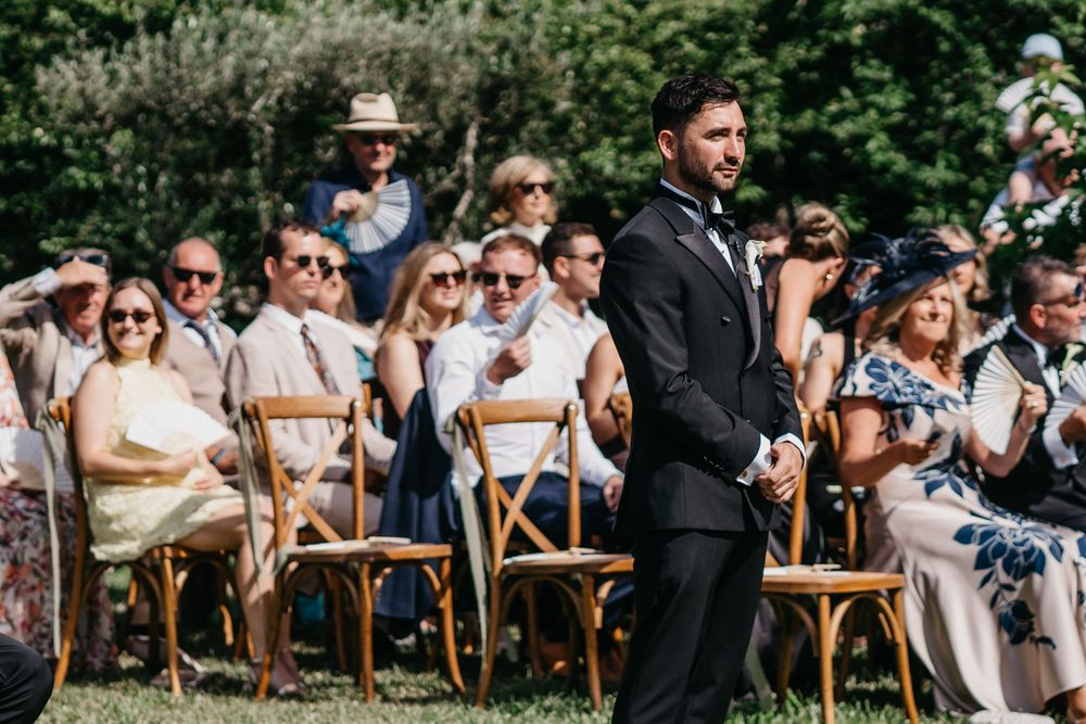 Groom in black tuxedo stands at outdoor wedding ceremony with seated guests in summer attire on wooden chairs.