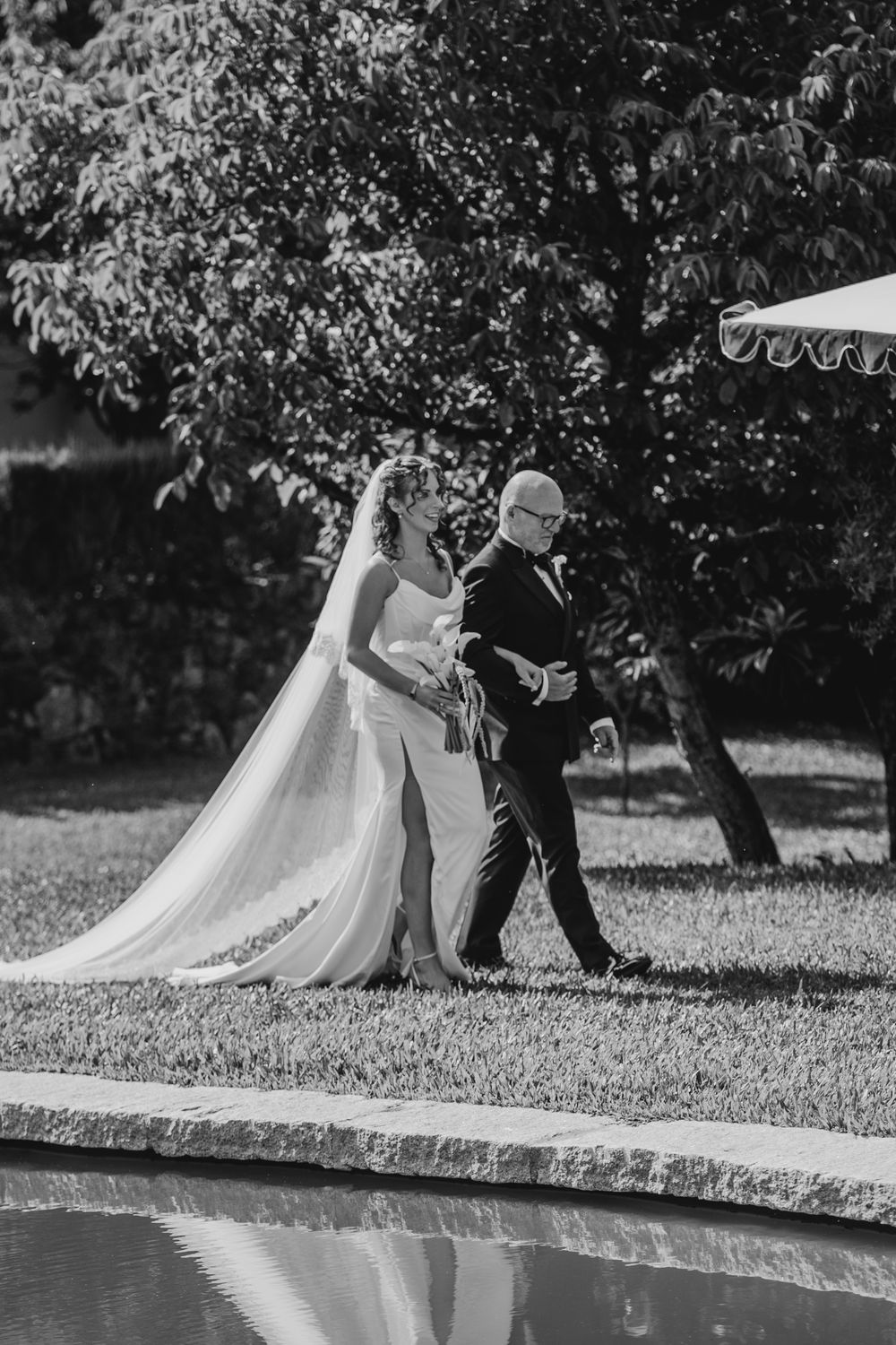 Black and white photo of a bride in a long veil walking with an older man in a suit beside a reflective pool.