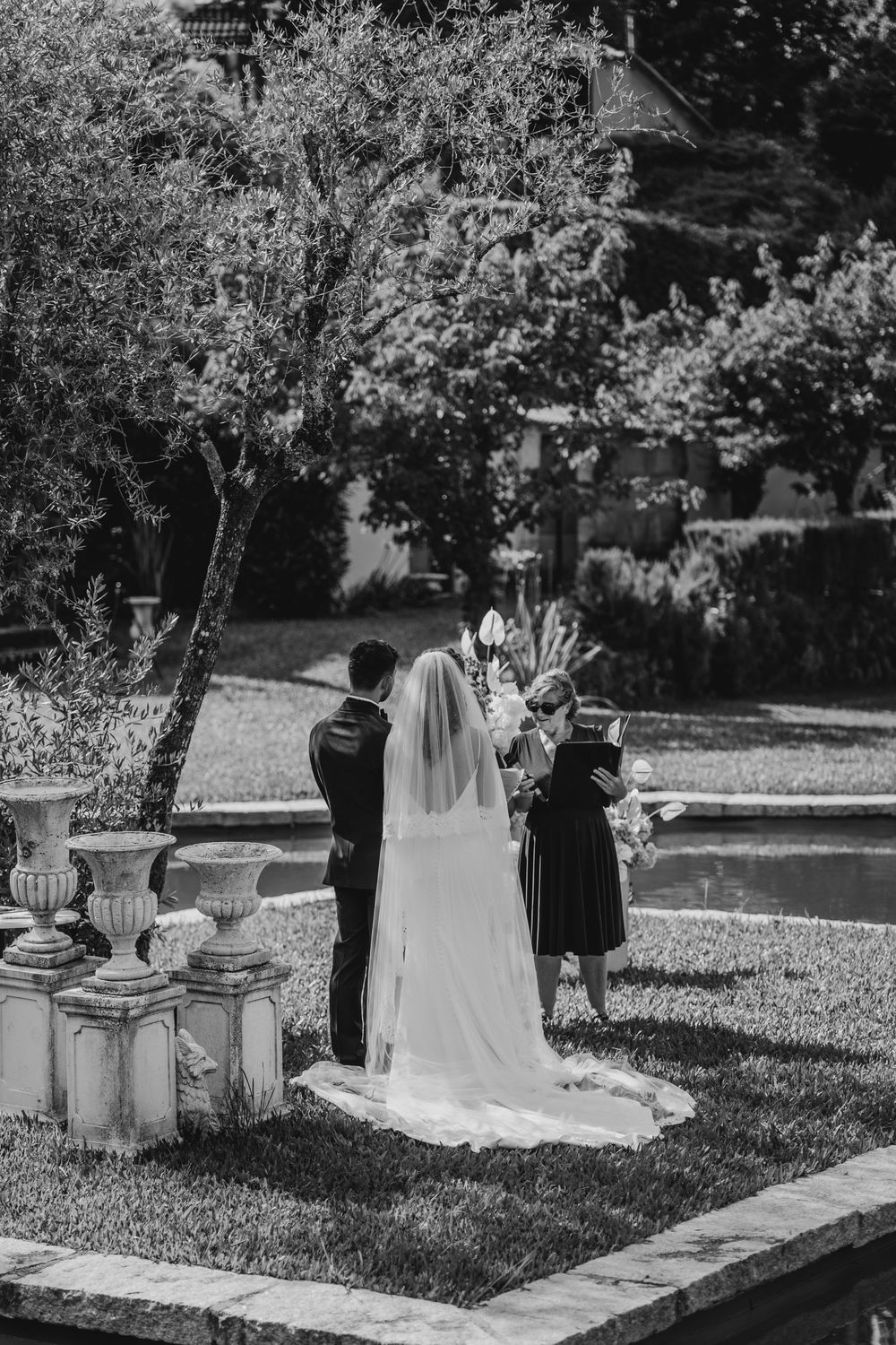 Black and white photo of a bride and groom during an outdoor garden wedding ceremony with lush trees and stone urns.