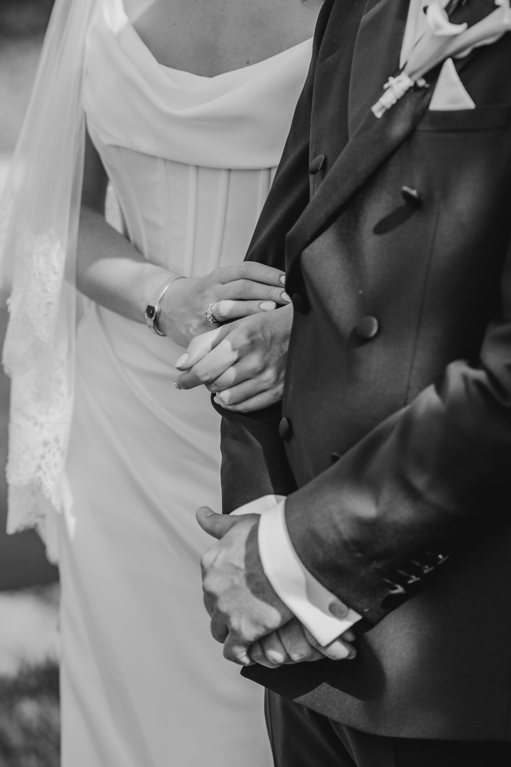 Black and white photo of bride and groom holding hands, showcasing wedding rings and elegant attire.