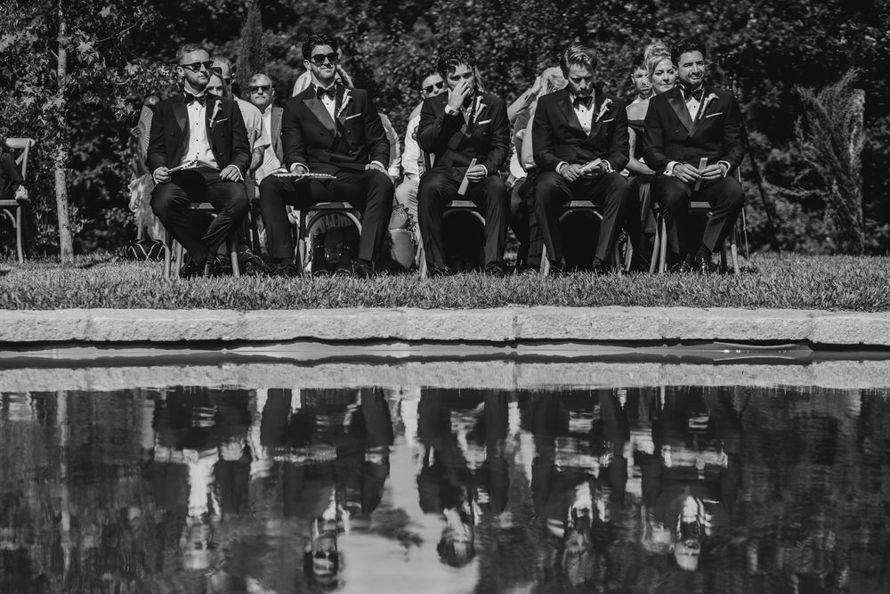 Black and white photo of groomsmen in tuxedos seated by a reflective pool at an outdoor wedding.