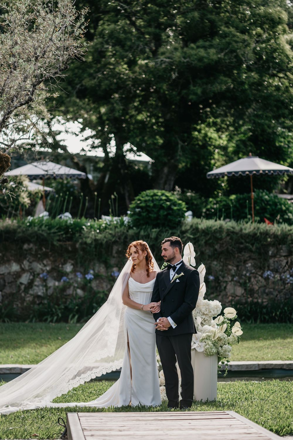 Bride in white gown with veil and groom in black tuxedo stand together during outdoor wedding ceremony.