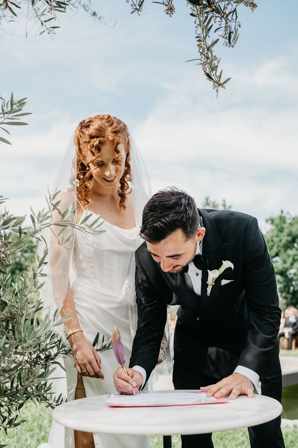 Bride and groom signing wedding registry outdoors, bride in white gown with veil, groom in black suit with boutonniere.