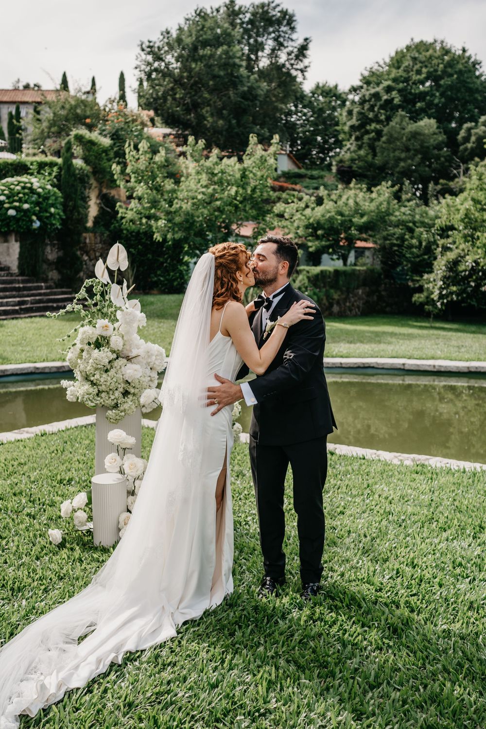 Bride and groom share first kiss at outdoor wedding ceremony by pond, surrounded by white floral arrangements.