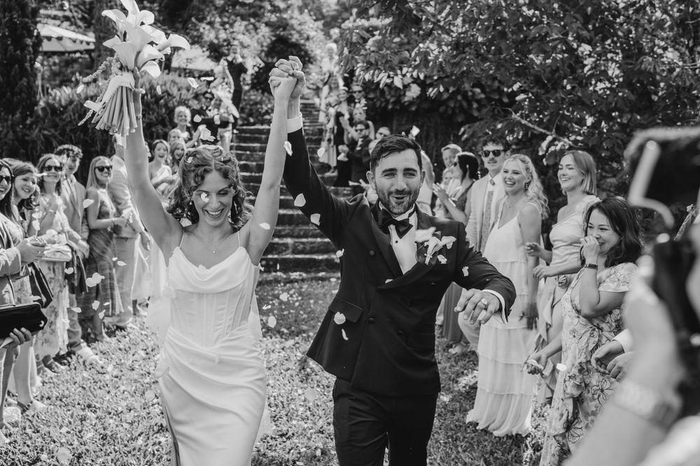 Black and white photo of a joyful newlywed couple raising hands while guests celebrate their wedding ceremony exit.