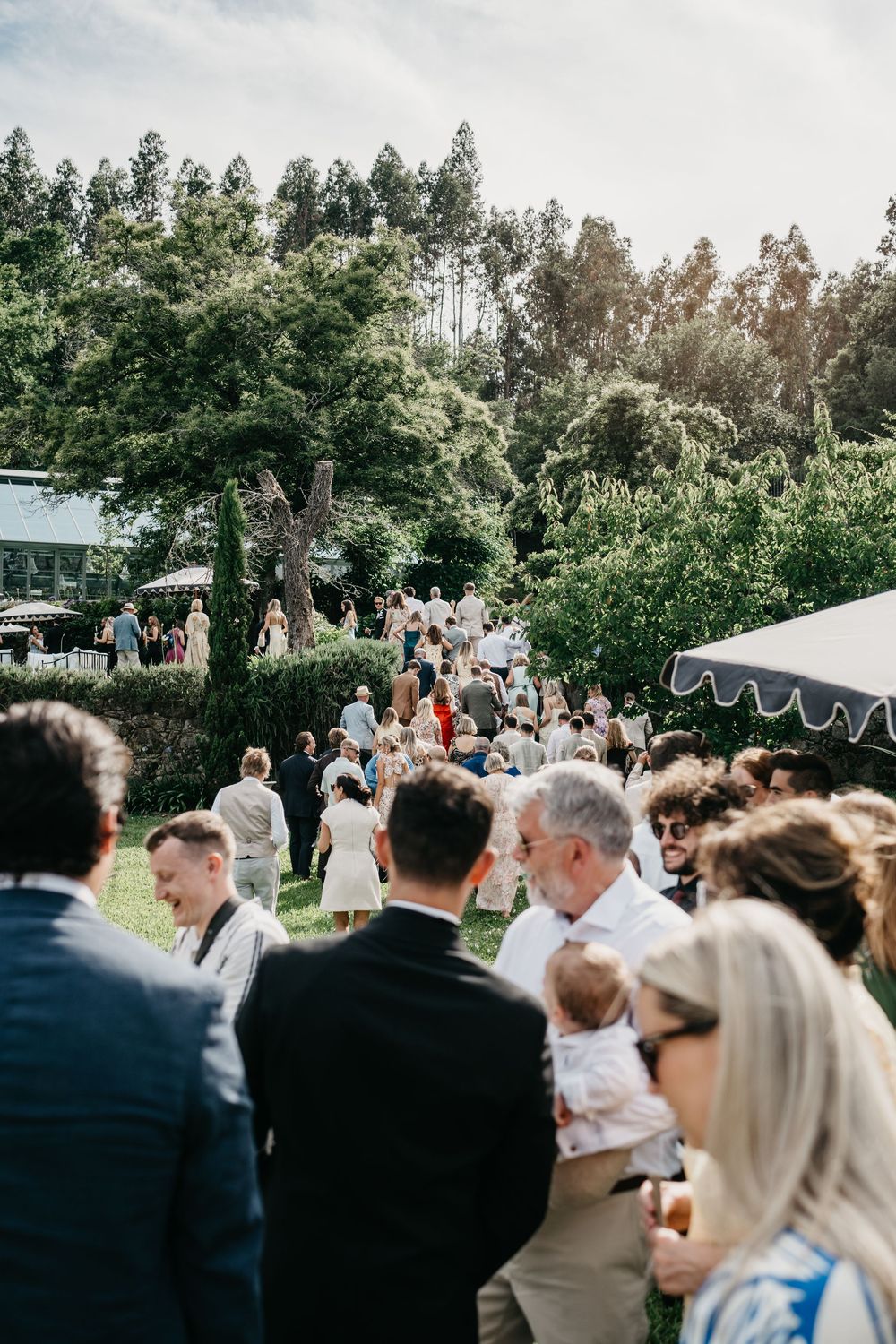 Guests mingling outdoors at a garden wedding reception with lush green trees and a white marquee tent in the background.