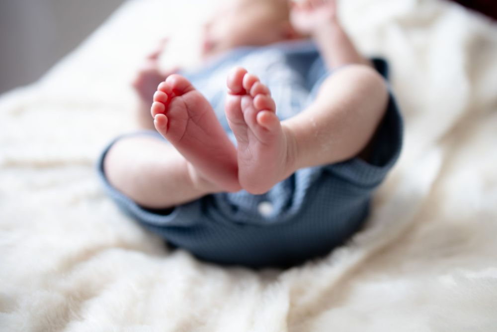 Close-up of adorable newborn baby feet and toes resting on a soft white blanket in blue pants.