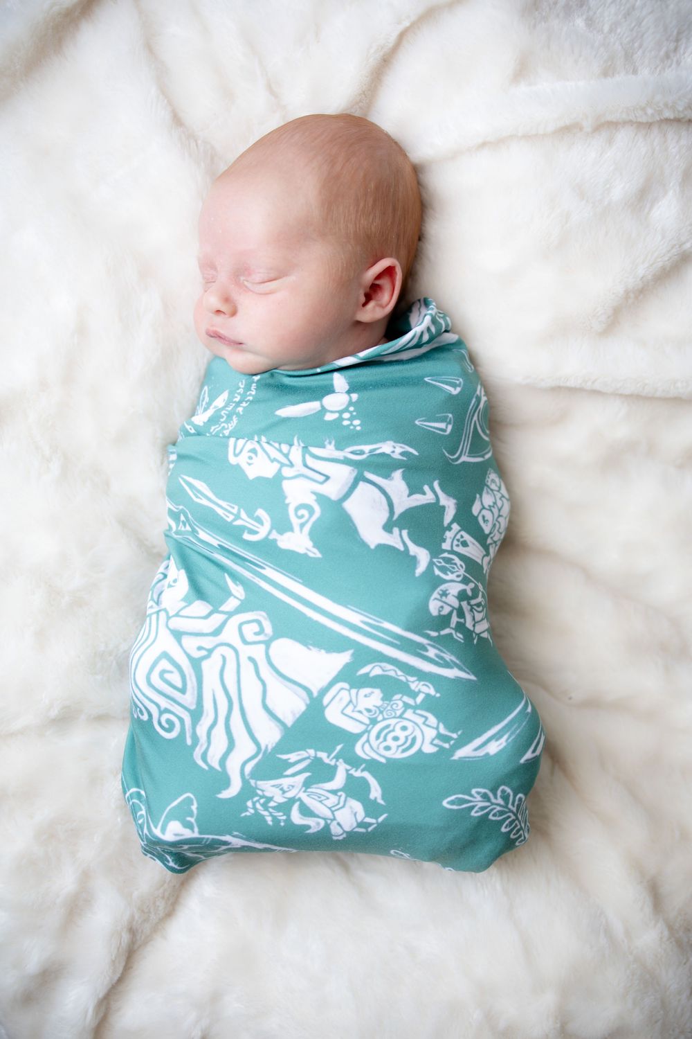 Newborn baby swaddled in a teal blanket with white patterns, sleeping peacefully on a white fluffy surface.