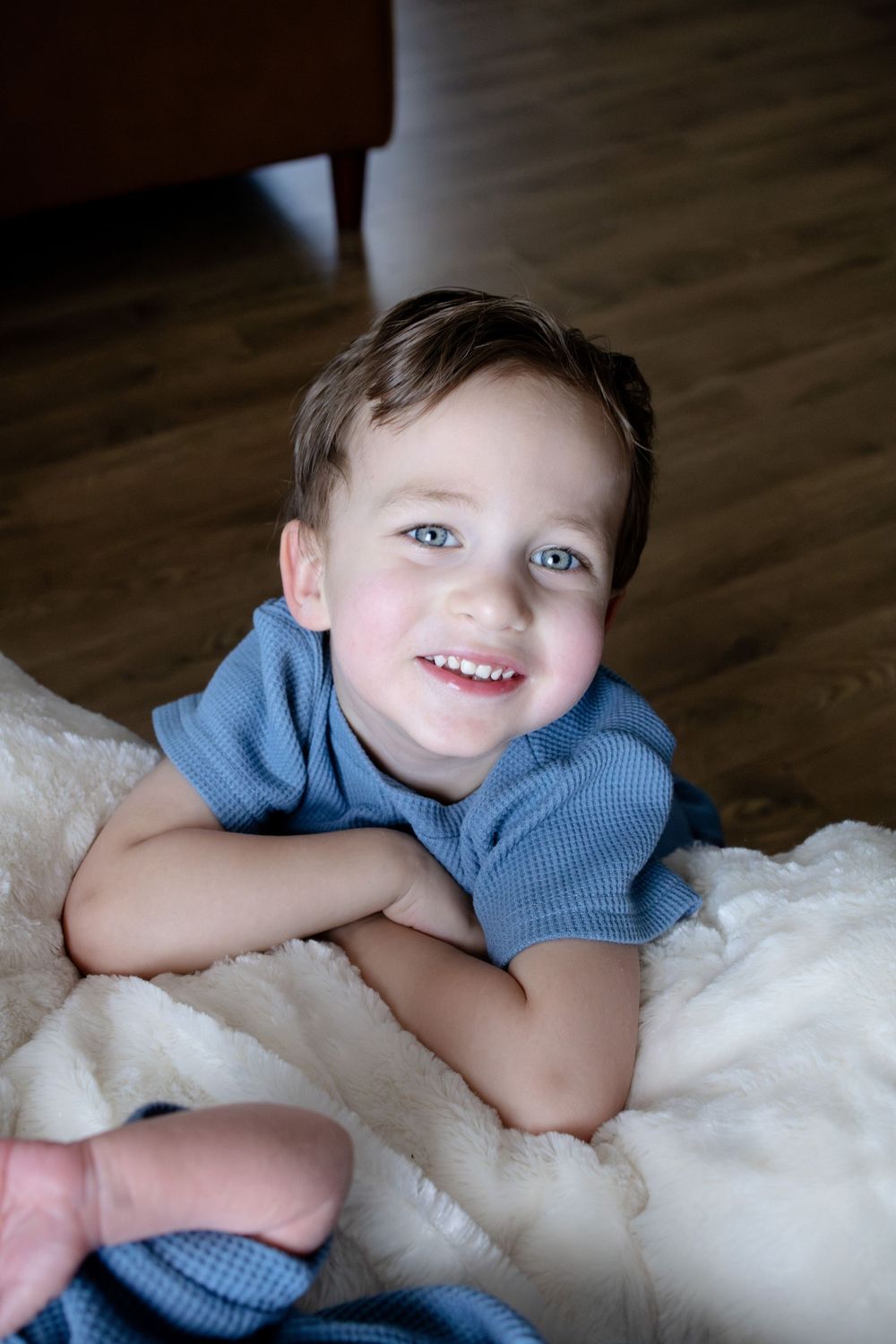 A smiling young boy with blue eyes lies on a fluffy white blanket wearing a blue shirt on a wood floor.