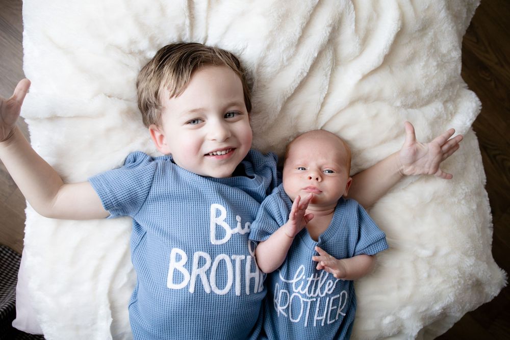 Big brother and little brother newborn boys wearing matching blue shirts lying on white fluffy blanket.