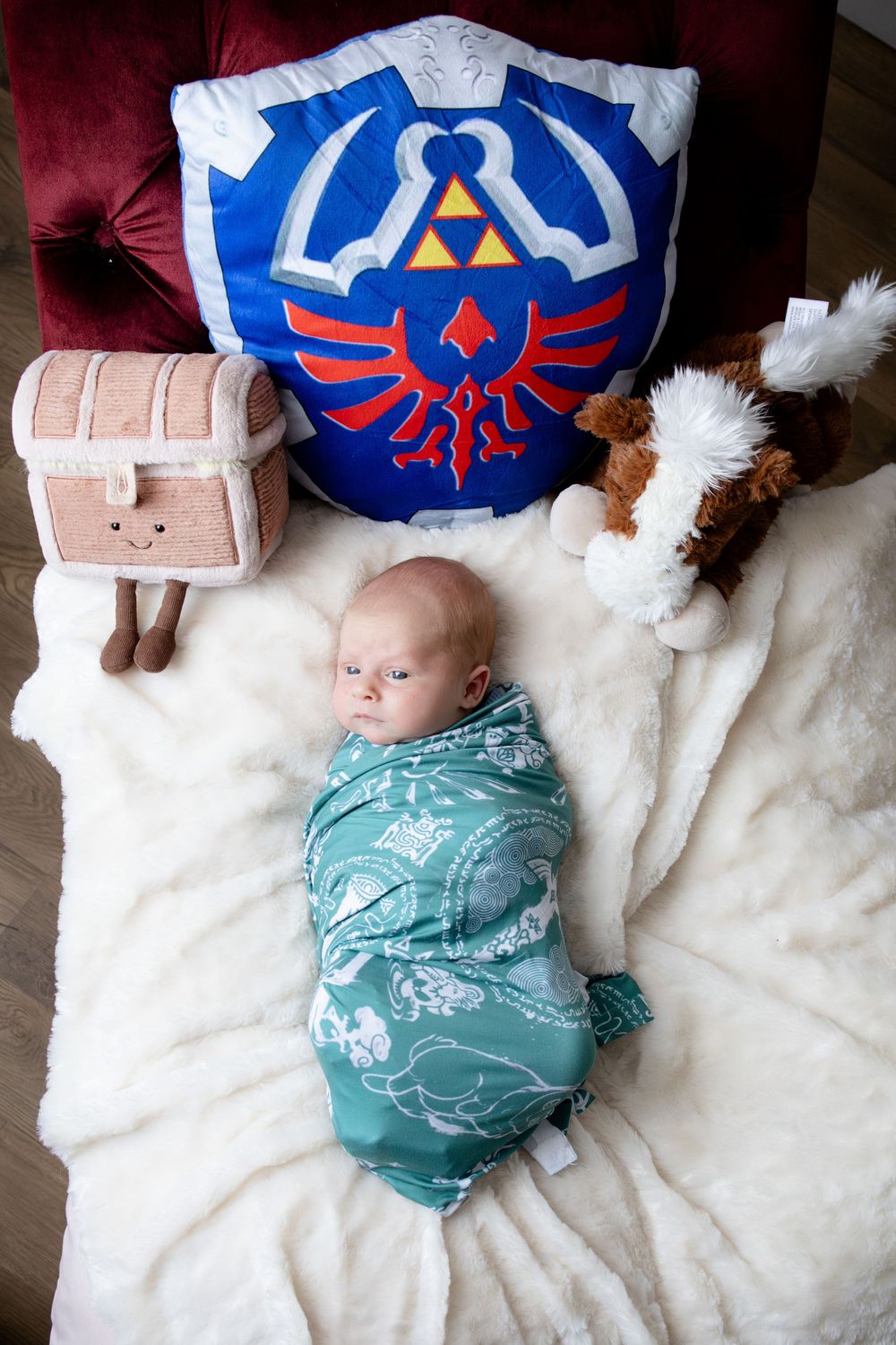 Baby swaddled in teal blanket surrounded by Legend of Zelda shield pillow, treasure chest plush, and horse stuffed animal.
