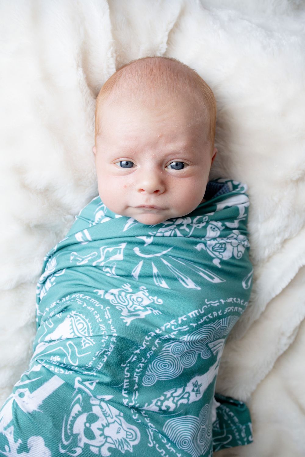 Newborn baby swaddled in a teal and white patterned blanket lying on a white fluffy surface.